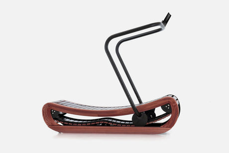 Side view of a curved manual treadmill with a dark reddish-brown wood frame, black slats, and a raised black handlebar